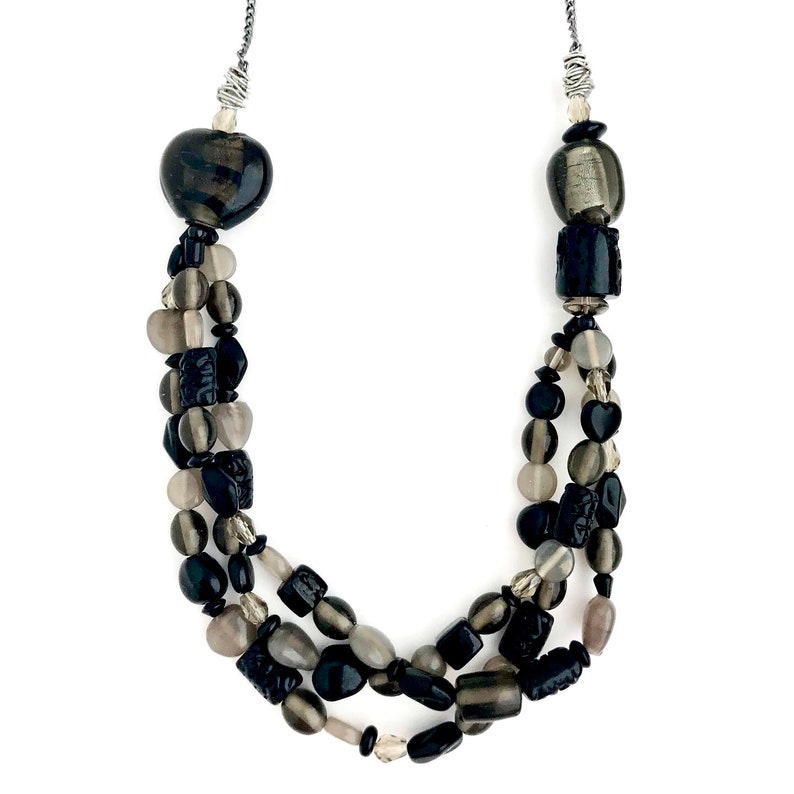 May include: A multi-strand necklace with black and gray beads. The necklace features a heart-shaped bead at the top and a variety of other shapes, including round, oval, and square beads.