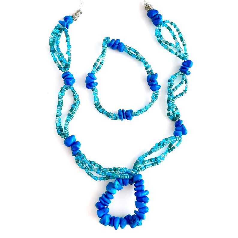 May include: A blue and turquoise beaded necklace and bracelet set. The necklace has a pendant in the shape of a circle. The bracelet is a simple band.