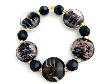 May include: A black and white bracelet with gold accents. The bracelet features round glass beads with abstract patterns and black faceted beads.