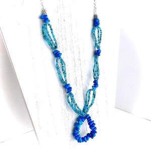 May include: A blue and turquoise beaded necklace with a silver clasp. The necklace is made of small, round beads and larger, irregular shaped beads. The necklace is displayed on a white background.