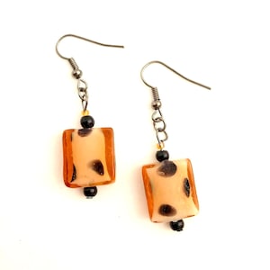 Puede incluir: Un par de pendientes colgantes con una cuenta cuadrada translúcida de color ámbar con manchas negras. Las cuentas están unidas a cadenas y ganchos plateados.
