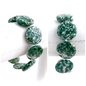 May include: A bracelet featuring large, round, green and white speckled beads. The beads are strung together with a clear string, creating a natural and earthy aesthetic. The bracelet is displayed against a white background, highlighting the colors and textures.