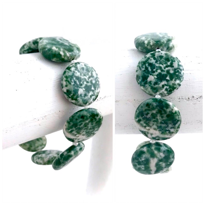 May include: A bracelet featuring large, round, green and white speckled beads. The beads are strung together with a clear string, creating a natural and earthy aesthetic. The bracelet is displayed against a white background, highlighting the colors and textures.