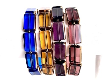 May include: A multi-strand bracelet featuring rectangular beads in shades of blue, amber, purple, and pink. The beads are strung together with gold-toned metal accents. The bracelet is displayed against a white background.