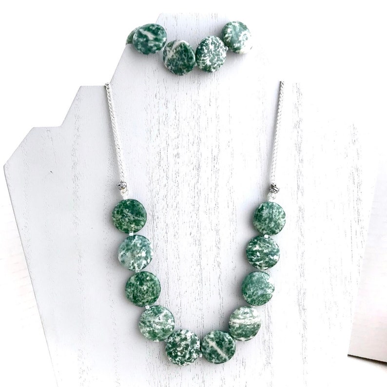 May include: A necklace and bracelet set featuring large, round beads with a speckled green and white pattern. The necklace hangs on a white cord, and the bracelet is displayed above. The jewelry is presented on a white display stand.
