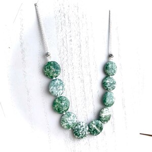 May include: A necklace featuring large, round, green and white speckled beads. The beads are strung on a silver-colored chain. The necklace is displayed on a white surface. The beads have a natural, organic appearance.