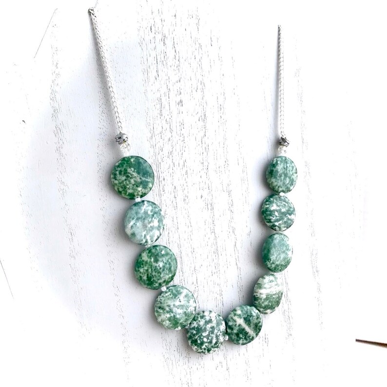 May include: A necklace featuring large, round, green and white speckled beads. The beads are strung on a silver-colored chain. The necklace is displayed on a white surface. The beads have a natural, organic appearance.