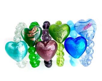 May include: A bracelet made of glass beads in various colors, including green, blue, and purple. The beads are shaped like hearts and have a metallic finish.
