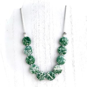 May include: A necklace featuring large, round, green and white speckled beads. The beads are strung on a silver-colored chain. The necklace is displayed against a white background.