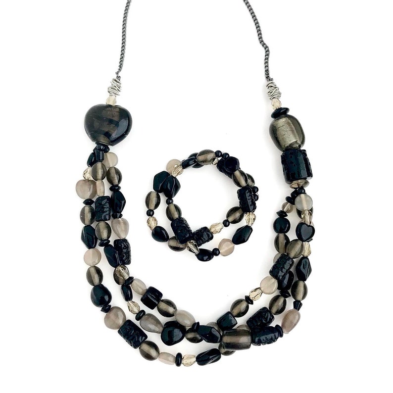 May include: A black and gray beaded necklace and bracelet set. The necklace is a triple strand of beads with a heart-shaped pendant. The bracelet is a single strand of beads.