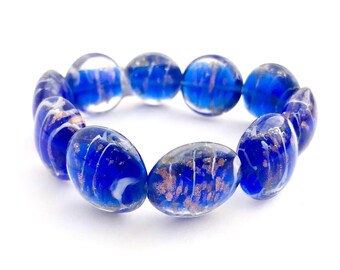 Könnte beinhalten: Ein blaues Glasarmband mit weißen und goldenen Sprenkeln. Das Armband besteht aus ovalen Perlen.