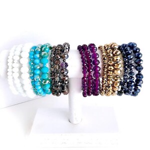 May include: A set of eight beaded bracelets in various colors, including white, turquoise, brown, purple, gold, and black. The bracelets are stacked on a white display stand.