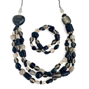 May include: A black and gray beaded necklace and bracelet set. The necklace is a triple strand of black and gray beads, with a heart-shaped pendant at the top. The bracelet is a single strand of black and gray beads.