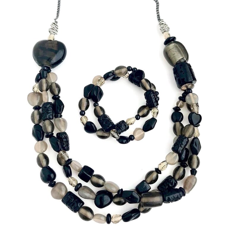 May include: A black and gray beaded necklace and bracelet set. The necklace is a triple strand of black and gray beads, with a heart-shaped pendant at the top. The bracelet is a single strand of black and gray beads.