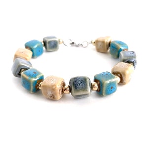 Puede incluir: Un brazalete hecho de cuentas cuadradas de cerámica en tonos azules, beige y grises. Las cuentas están separadas por pequeñas cuentas doradas.