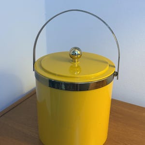 Pode incluir: Um balde de gelo cilíndrico amarelo brilhante com acabamento liso. Apresenta uma borda de metal prateado polido, uma alça oscilante prateada correspondente e uma tampa com um pequeno puxador esférico de cor dourada.