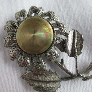 May include: A silver tone brooch in the shape of a flower with a round, iridescent, gold-toned center.