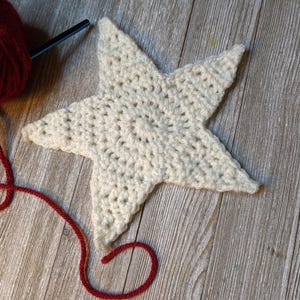 May include: A cream-colored, crocheted star lies on a wooden surface, with a ball of red yarn and a crochet hook in the upper left corner. The star is the focal point, showcasing the texture of the handmade craft. The yarn's red color provides a contrast to the star.