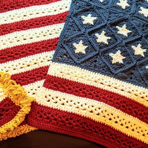 Könnte beinhalten: Gehäkelte Decke und herzförmige Dekoration im Stil der amerikanischen Flagge. Die Decke hat rote und weiße Streifen mit einem blauen Quadrat mit weißen Sternen. Die herzförmige Dekoration hat ein ähnliches Design.