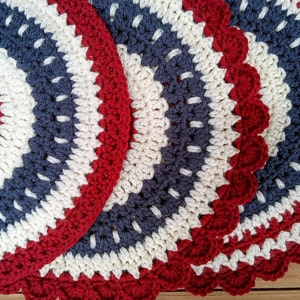 May include: A collection of handmade crochet placemats. Each placemat features a circular design with concentric rings of red, white, and navy blue yarn. The edges have a scalloped trim.