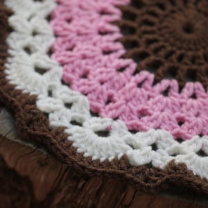 May include: A close-up of a crocheted round doily with brown, white, and pink yarn. The doily is on a wooden surface.