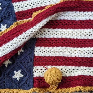 Op de afbeelding: Een handgemaakte gehaakte deken in de stijl van de Amerikaanse vlag. De deken heeft afwisselend rode en witte strepen, een blauw veld met witte sterren en een goudkleurige geschulpte rand. Een bol wol en een haaknaald zijn ook zichtbaar.