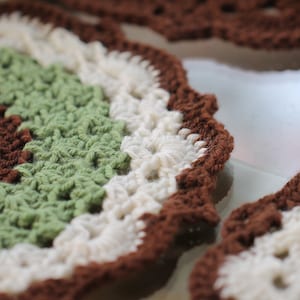 May include: Close-up of a crocheted trivet with brown, white, and green yarn. The trivet is circular and has a decorative border.