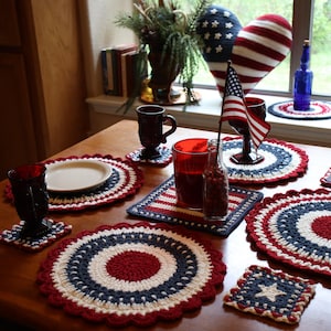 Pode incluir: Uma coleção de jogos americanos e porta-copos de crochê nas cores vermelho, branco e azul com designs circulares e quadrados. O conjunto inclui uma pequena bandeira americana, copos vermelho-escuros e uma almofada em forma de coração.