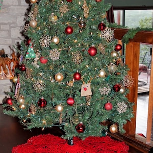 May include: A red crocheted Christmas tree skirt with a white lace trim. The skirt is placed under a decorated Christmas tree with red and gold ornaments.