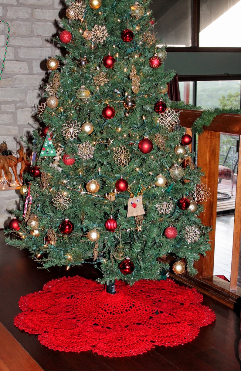 May include: A red crocheted Christmas tree skirt with a white lace trim. The skirt is placed under a decorated Christmas tree with red and gold ornaments.