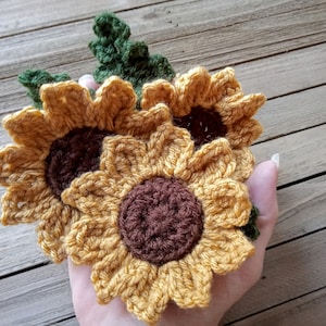 May include: Three crocheted sunflowers with brown centers and green leaves. The sunflowers are yellow and brown.