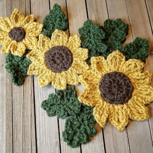 May include: Three crocheted sunflowers with brown centers and yellow petals, surrounded by five green crocheted leaves. The flowers and leaves are arranged in a heart shape on a wooden surface.