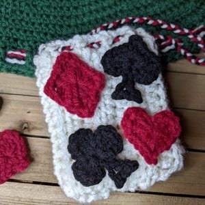 May include: A crocheted playing card design with red diamond, heart, and black spade and club symbols on a white background. The card is on a wooden surface with a green crocheted border.