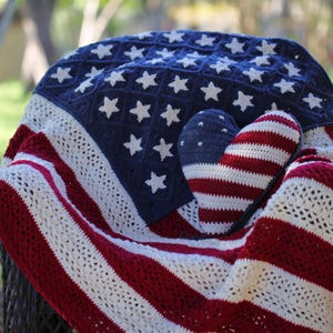 Puede incluir: Una manta y un cojín en forma de corazón de ganchillo en rojo, blanco y azul, que se asemejan a la bandera estadounidense. La manta presenta estrellas blancas sobre un campo azul y rayas rojas y blancas. El cojín es rayado.