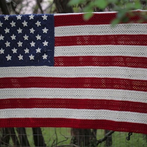 Puede incluir: Una manta de ganchillo que representa la bandera estadounidense, con un campo azul de estrellas blancas y rayas rojas y blancas alternas. La manta está colgada sobre una valla de madera, mostrando el diseño patriótico.