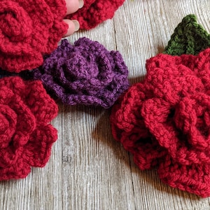 Puede incluir: Una colección de flores de ganchillo hechas a mano en varios colores. La imagen presenta varias rosas rojas, una rosa morada y hojas verdes. Las flores están dispuestas sobre una superficie de madera, mostrando la textura y el detalle del trabajo de ganchillo.