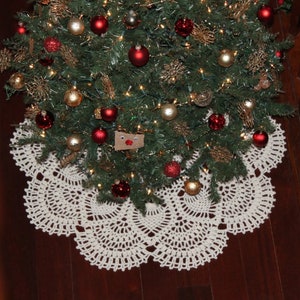 May include: A white crocheted Christmas tree skirt with a scalloped edge. The skirt is placed on a wooden floor under a decorated Christmas tree.