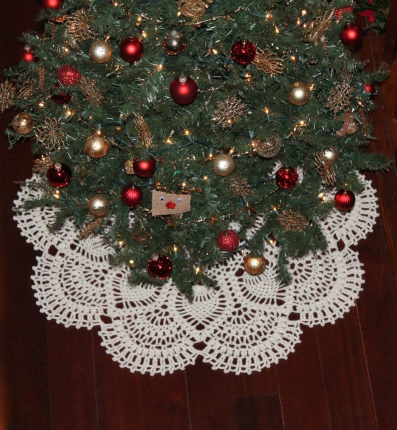 May include: A white crocheted Christmas tree skirt with a scalloped edge. The skirt is placed on a wooden floor under a decorated Christmas tree.