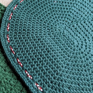 May include: Close-up of a handmade, dark teal crochet coaster with a rounded edge. The edge is detailed with a red and white stitched pattern. The coaster is part of a set, with a portion of a second, similar coaster visible.