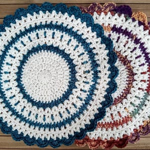 May include: Two handmade crochet doilies. One is white with teal blue accents and a scalloped edge. The other is a mix of colours including purple, brown, and orange. The doilies are circular and sit on a wooden surface.