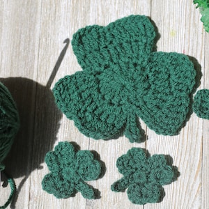May include: Four green crocheted shamrocks of varying sizes, with a ball of green yarn and a crochet hook.