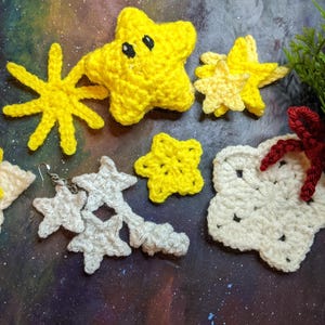 Pode incluir: Uma coleção de estrelas de crochê em vários tamanhos e cores, incluindo amarelo, branco e creme. As estrelas estão dispostas sobre um fundo azul escuro com um padrão de galáxia.