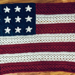 Pode incluir: Um cobertor de crochê feito à mão com o design da bandeira americana. Possui um quadrado azul marinho com estrelas brancas e listras vermelhas e brancas alternadas. O trabalho de crochê é visível, adicionando textura.