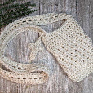 May include: A crocheted cream-colored bag with a long strap and a small crocheted cross charm. The bag is made with a simple stitch pattern and has a slightly textured surface.