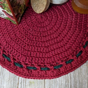May include: A circular, hand-crocheted trivet in a rich red colour, featuring a decorative border of dark green stitching. The trivet is made from thick yarn and is designed to protect surfaces from hot items. Other items are partially visible.