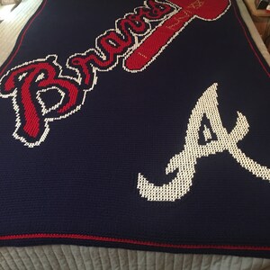May include: A navy blue crocheted blanket with the Atlanta Braves logo in red and white. The logo includes the team's name "Braves" and the letter "A".