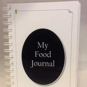 May include: A white spiral bound journal with a black circle on the cover. The circle has the text "My Food Journal" in white.