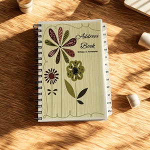 May include: Spiral-bound address book with a light green cover featuring floral designs in brown, green, and black. The words "Address Book Birthdays & Anniversaries" are printed on the cover. The book has alphabetical tabs.