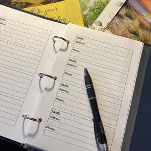 May include: An open address book with lined pages and metal ring binders. A black pen rests on the page, ready to write. The pages are labeled with "NAME: Address: Phone:" and a greeting card is visible in the background.