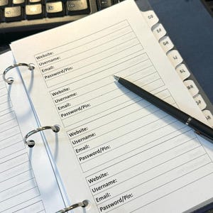 May include: An open binder with lined pages for recording website, username, email, and password/PIN information. A black pen rests on the page. The binder is open on a desk with a keyboard and tabbed dividers.